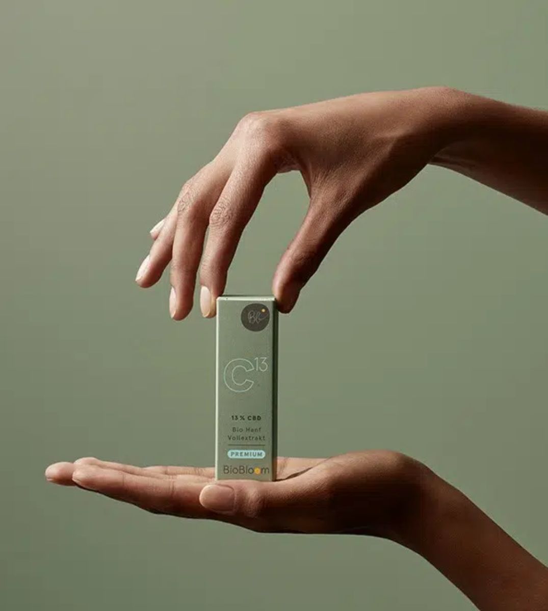 Hände präsentieren elegant die Verpackung von BioBloom C13 mit 13% CBD Vollextrakt auf einem einfarbigen olivgrünen Hintergrund
