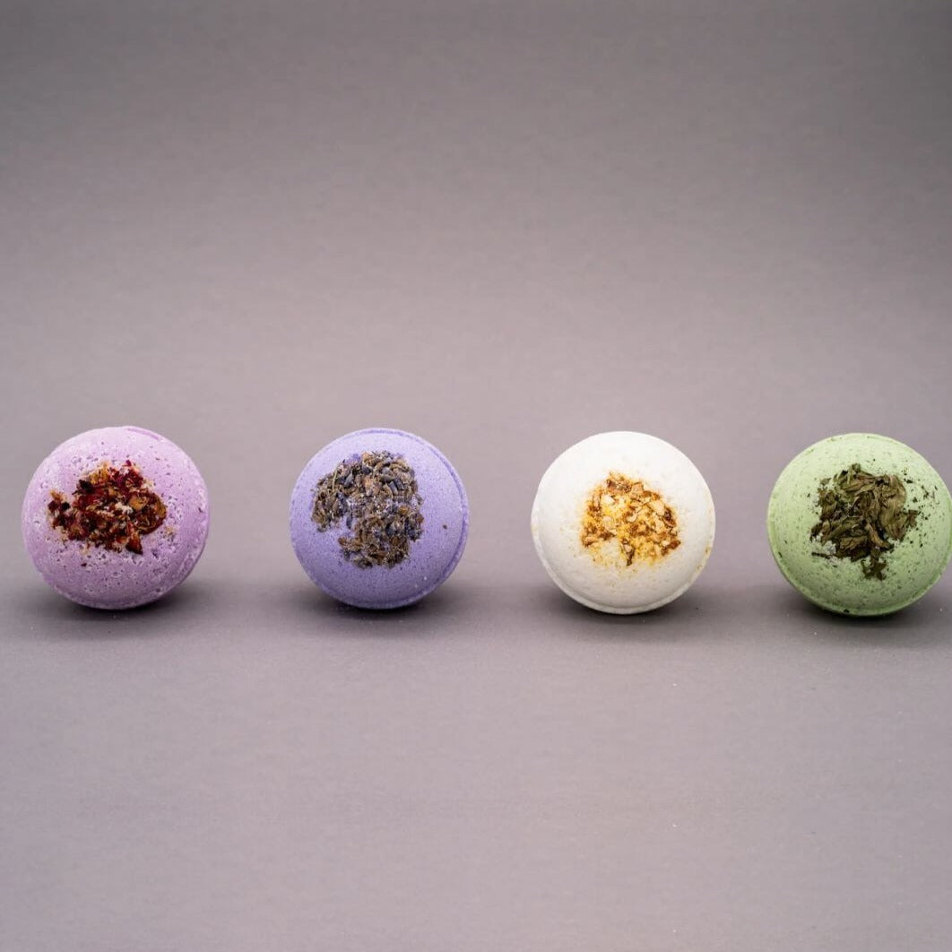 Vier verschiedene CBD-Badebomben mit Blütenblätter-Toppings in Lila, Blau, Weiß und Grün, präsentiert auf einem neutralen grauen Hintergrund für entspannende Wellness-Baderituale