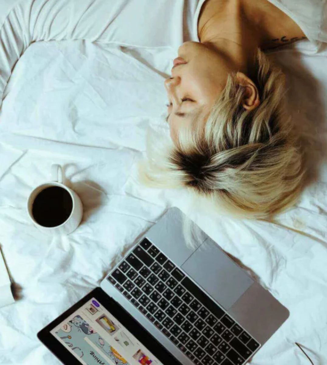 Entspannte Frau liegt im Bett mit einem Laptop, einer Tasse Kaffee und einem Notizbuch, Konzept für Home-Office und Entspannung.