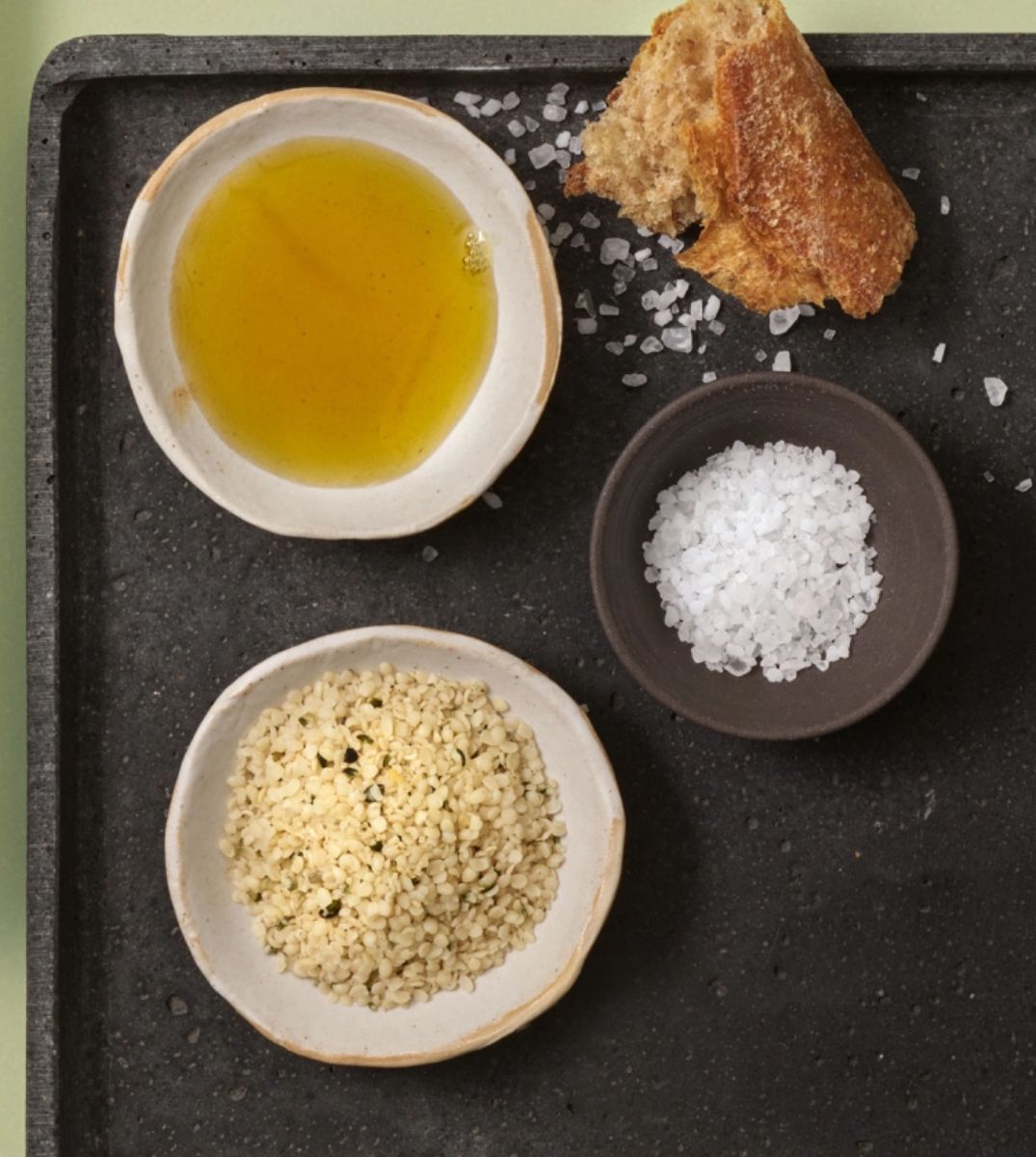 Olivenöl, grobes Salz und Hanfsamen in Schalen auf Schieferplatte mit Baguettestück, für eine gesunde Mahlzeit arrangiert
