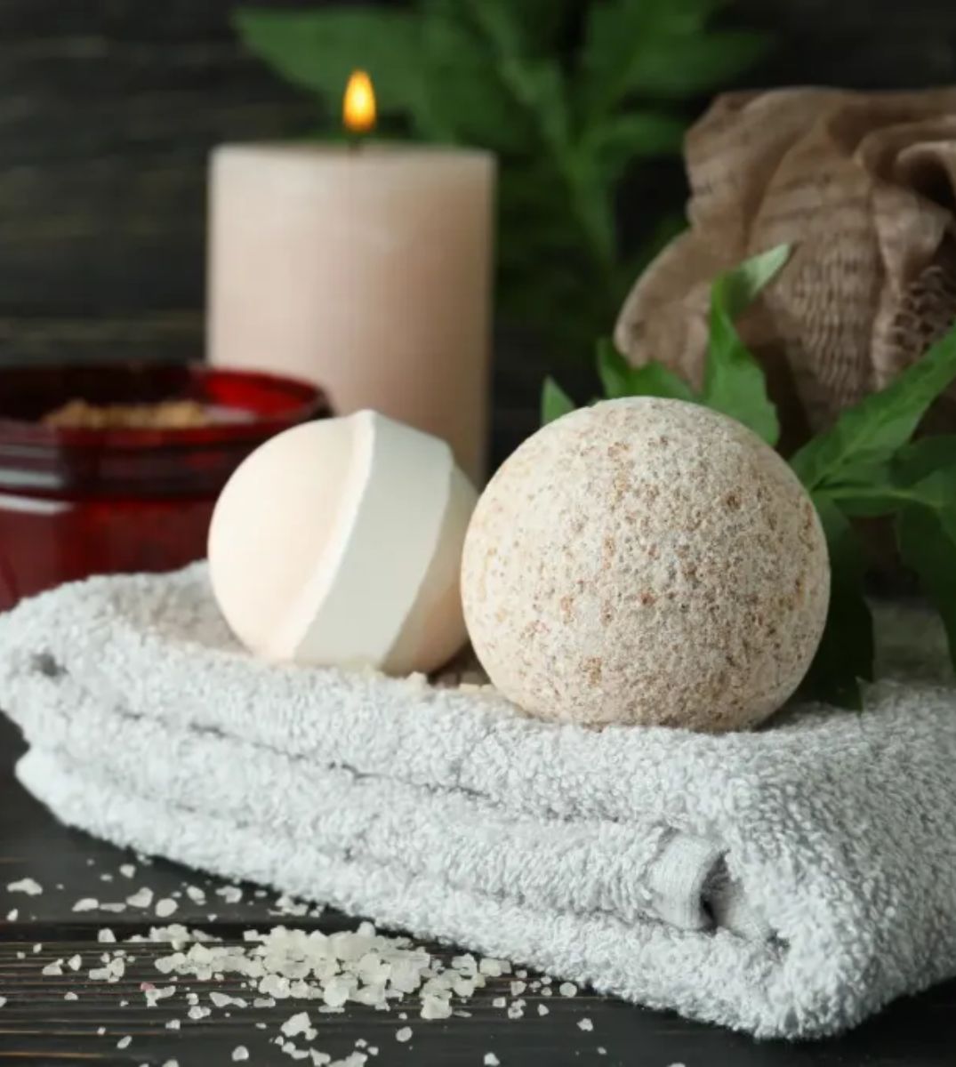 Stillleben-Anordnung für Wellness und Entspannung mit Badebomben auf einem flauschigen Handtuch, begleitet von einer brennenden Kerze und einem Glas Hautpflegeprodukt auf einem dunklen Holzhintergrund.